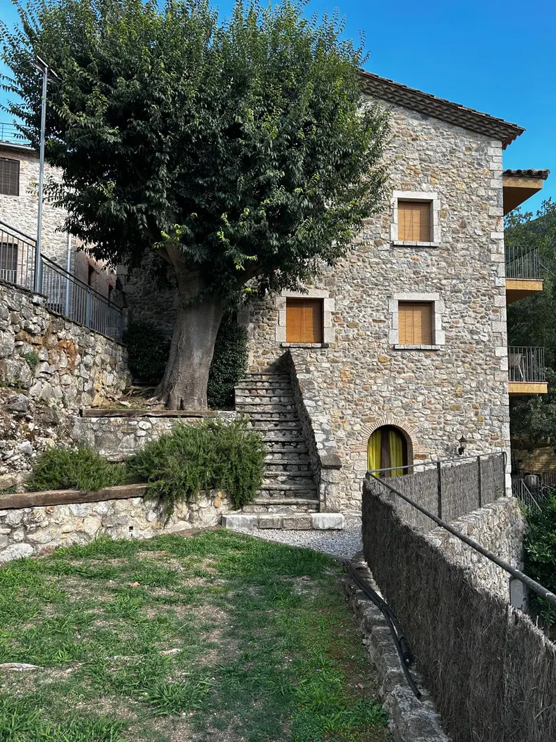 Façana de pedra de Can Martinot amb l’arbre, les escales d’accés i la gespa